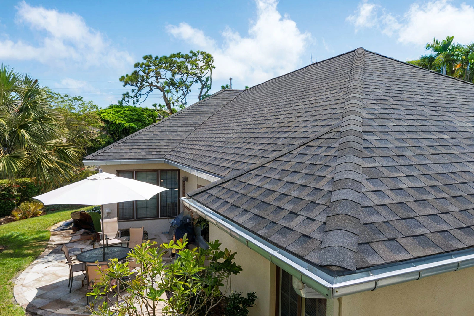 A sloped roof covered with dark shingles overlooks a patio with seating and greenery.